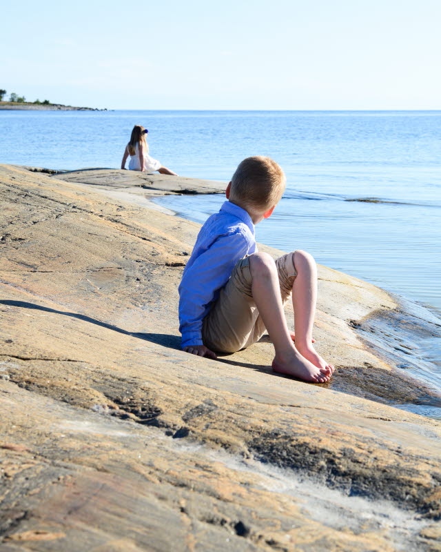 Barn sitter en bit ifrån varandra på en klippa vid havet. Barnet närmast vänder sig om