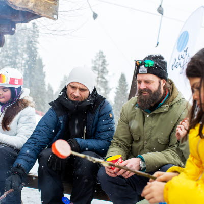 Fyra personer sitter och grillar korv i ett vindskydd vid en slalombacke.