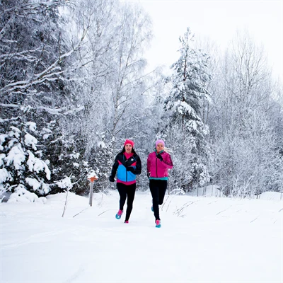 Två kvinnor löper i skoterspår. Marken, träden och buskarna har snö på sig. KVinnonra är klädda i starka kontrastfärger.