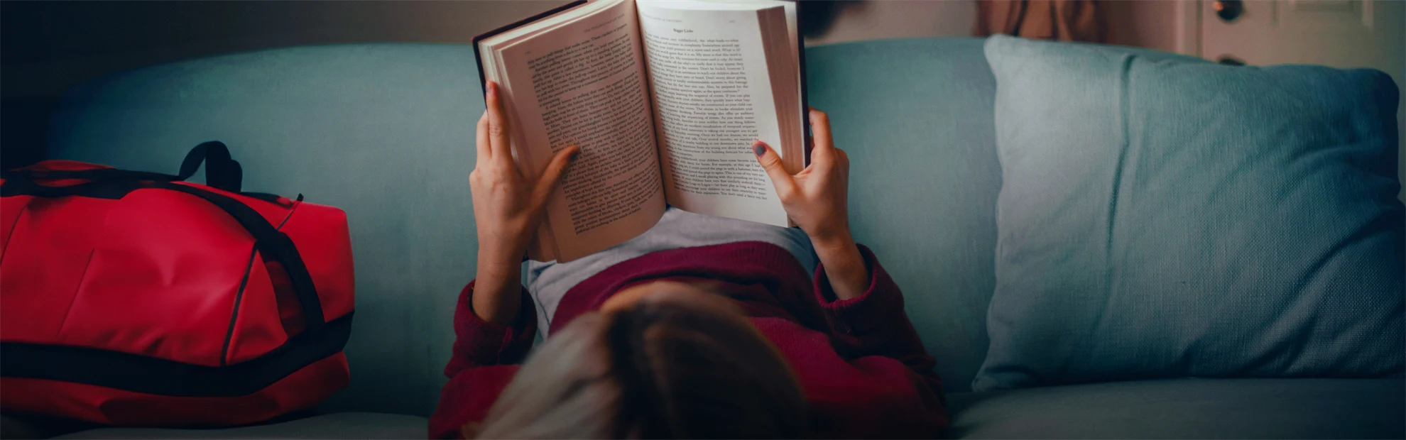 En person ligger med fötterna på ryggstödet och huvudet på sittkudden på en soffa och läser en bok.