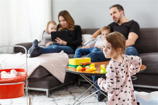 En familj sitter samlade i ett vardagsrum. Två vuxna och två barn sitter i soffan, medan ett tredje barn står framför soffbordet. Det finns leksaker i rummet och några av dem tittar på en surfplatta.