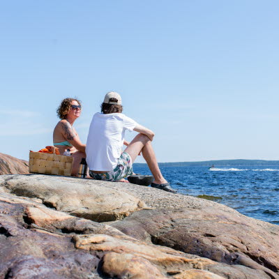 Två vuxna personer sitter med en picknickkorg på klippor vid ett hav och i bakgrunden syns en segelbåt.