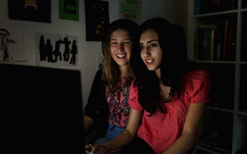 Två unga tjejer sitter i mörkt rum och ser på en dataskärm som lyser upp deras ansikten. 