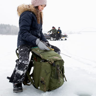 En ung tjej sitter och pimplar på isen med två personer som sitter på en skoter i bakgrunden.