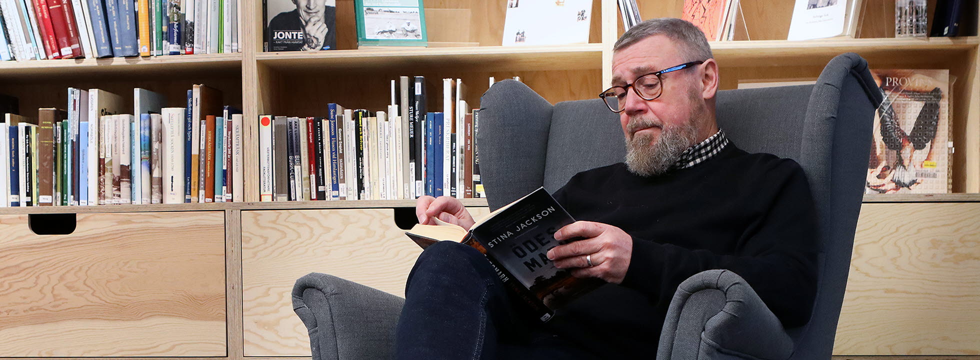 Jonas Lundqvist sitter i en fåtölj med hög rygg, han har benen i kors och läser en bok som han håller i knät.