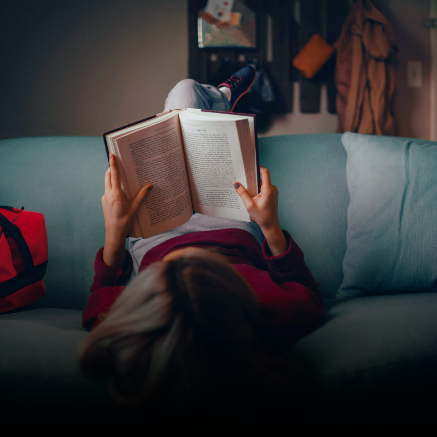Ungdom som ligger med fötterna över ryggstödet och huvudet ner mot golvet i en soffa och läser en bok. Bredvid i soffan ligger en träningsväska.