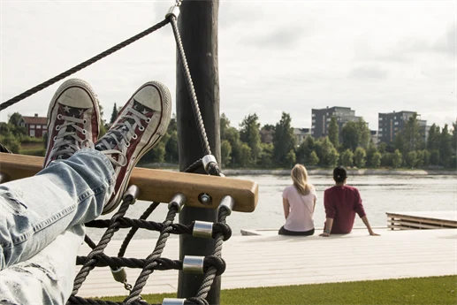 Människa ligger i hängmatta i älvsbrinken, med vy över älven, och människor som sitter på brygga.