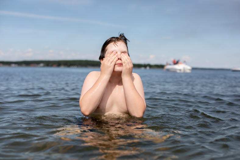 Ett barn badar i havet på sommaren med en båt som syns i bakgrunden.