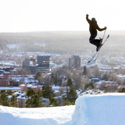 En person gör ett trick i ett hopp i vitbergsbacken och i bakgrunden ser man delar av Skellefteå stad.