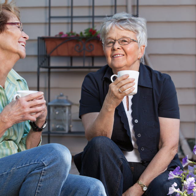 Två kvinnor sitter på en altantrapp och pratar, ler och dricker ur mugg