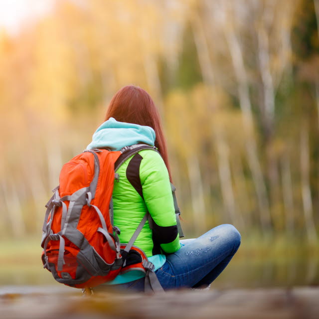 En person med ryggsäck sitter på en brygga och tittar mot skogen som är höstfärgad