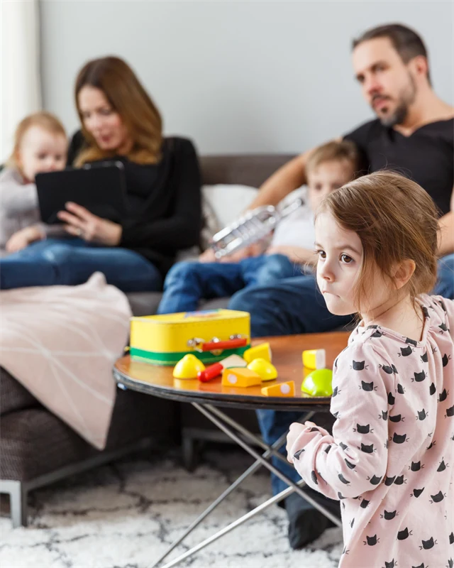 En familj sitter samlade i ett vardagsrum. Två vuxna och två barn sitter i soffan, medan ett tredje barn står framför soffbordet. Det finns leksaker i rummet och några av dem tittar på en surfplatta.