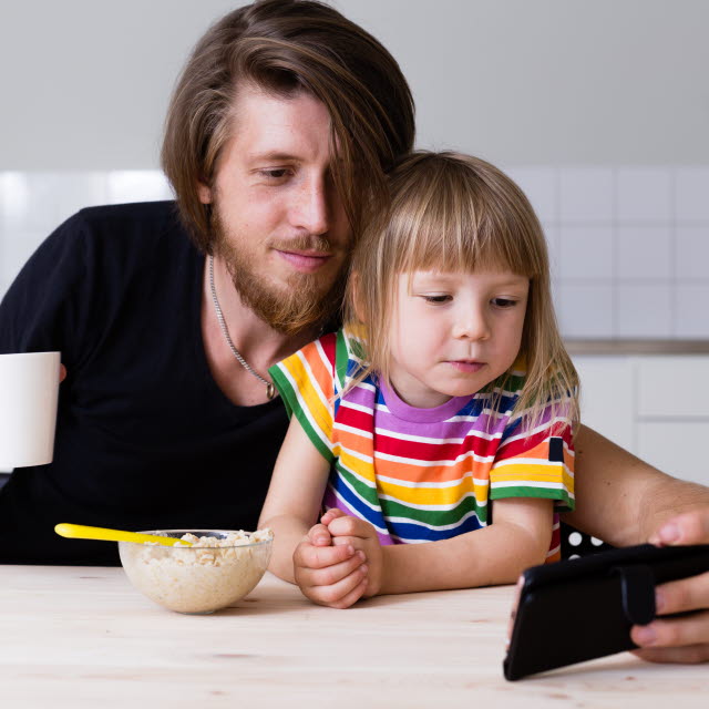 Ett barn sitter i en mans knä vid ett bord. Mannen håller i en kaffekopp och tillsammans tittar dom på mobilen. Barnet har en gröttallrik framför sig.