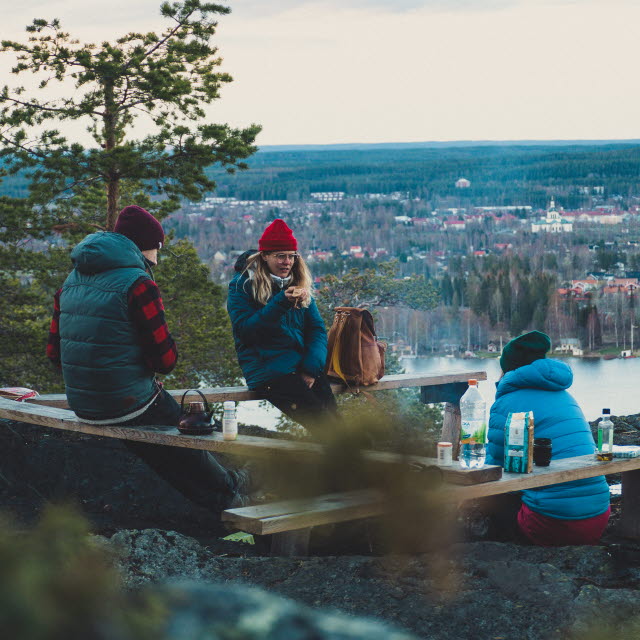 Tre personer sitter och fikar vid en brasa uppe på ett berg. I bakgrunden utsikt över Skellefteå.
