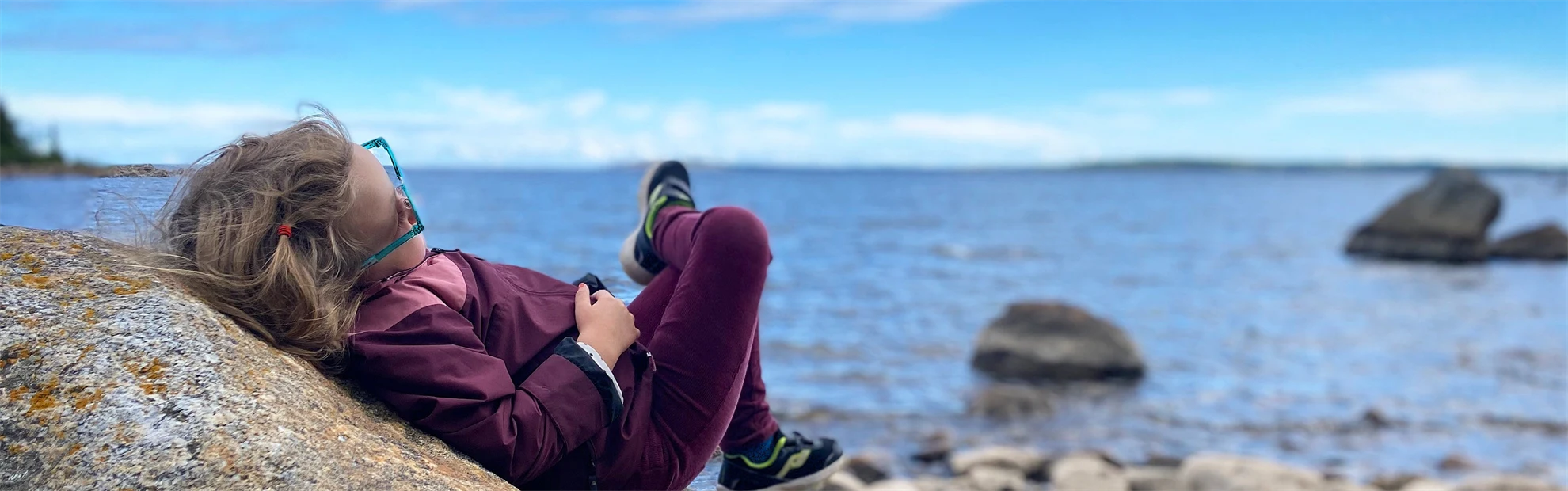 En kvinna i solglasögon ligger på klippor vid ett hav och läser en bok.