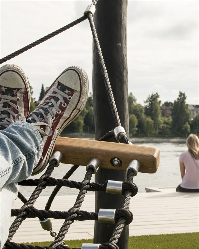 Människa ligger i hängmatta i älvsbrinken, med vy över älven, och människor som sitter på brygga.